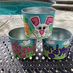 May include: Three metal pails painted with colorful designs. The top pail has a white bunny with pink ears and a carrot. The middle pail says "Kendra" in pink and purple. The bottom pail says "Jordyn" in purple and green.