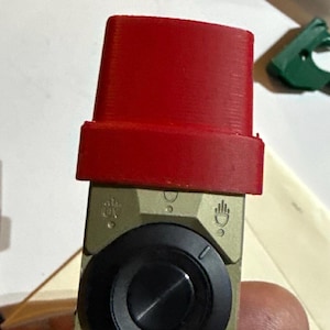 Könnte beinhalten: Ein hellbeiger Autoscheinwerferschalter mit einem zentralen schwarzen runden Drehknopf. Der Schalter hat eine rote Kunststoffabdeckung an der Oberseite. Eingravierte Symbole auf der beigen Oberfläche zeigen verschiedene Lichteinstellungen an, darunter ein Handsymbol.