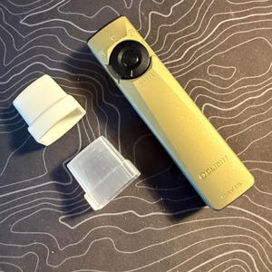 Könnte beinhalten: Eine khakifarbene Taschenlampe mit einem schwarzen Bedienrad und einer transparenten Kunststoffabdeckung. Zwei weiße und transparente Kunststoffzubehörteile liegen neben der Taschenlampe. Die Taschenlampe trägt die Marke "OLIGHT".