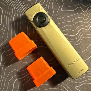 May include: A tan-colored flashlight with a black circular button and a series of indicator lights. Two bright orange rectangular accessories are positioned near the flashlight. The flashlight has the brand name "OLIGHT" printed on it.