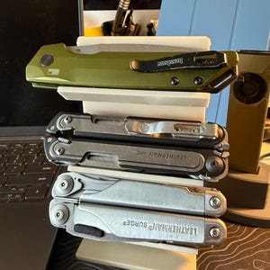May include: An assortment of multi-tools and a folding knife are displayed on a white stand. The top knife is green with a black clip and the word "Kershaw". Below are two Leatherman multi-tools, one with "Leatherman ARC" and the other with "Leatherman Surge" printed on them.