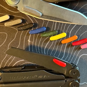 Pode incluir: Uma multi-ferramenta preta com uma lâmina parcialmente aberta e uma lâmina fechada, juntamente com vários pequenos acessórios cilíndricos coloridos. Os acessórios são em várias cores, incluindo branco, cinza, preto, azul, verde, amarelo, laranja, vermelho e rosa.