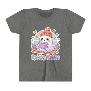 Peut inclure: Un t-shirt gris chiné foncé avec un motif représentant un petit fantôme blanc portant un bonnet orange, lisant un livre et assis sur une couverture entre deux tasses de café. Le design inclut un arc-en-ciel pastel, des cœurs, des étoiles et le texte Spooky Stories.