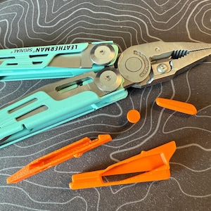 May include: A multi-tool with a teal handle and the text "LEATHERMAN SIGNAL" is open, revealing a blade. Several orange plastic accessories are scattered around the tool. The multi-tool is on a dark surface with a topographic pattern.