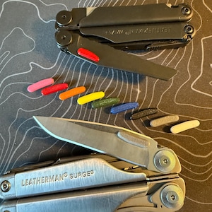 May include: Two multi-tools, one black and one silver, with various colored handle inserts. The black tool is partially open, revealing a red insert. The silver tool is labeled "LEATHERMAN SURGE". A variety of colored inserts are scattered nearby.