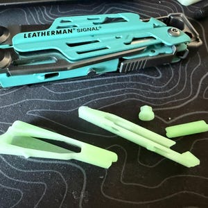 May include: A multi-tool with a teal handle, labeled "LEATHERMAN SIGNAL", is shown open, revealing various tools. Several lime green replacement parts are scattered nearby. The tools are on a dark surface with a topographic pattern.
