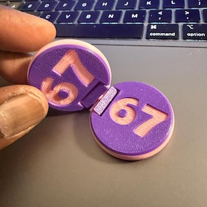 Könnte beinhalten: Zwei lila und pfirsichfarbene kreisförmige Objekte mit der Nummer "67" in Pfirsich. Die Objekte befinden sich auf einer Laptop-Tastatur. Ein Objekt ist teilweise vom anderen getrennt.