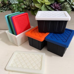 May include: A selection of small storage containers with lids in various colours. The containers are black with lids in white, orange, and blue. A stack of coloured lids in red, green, and blue are in a white container. A white lid with a diamond pattern is also present.