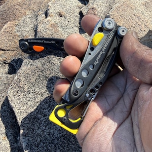 May include: Two black and silver multi-tools with yellow and orange accents are displayed. One is open, revealing a blade. The text "LEATHERMAN Skeletool CX" is visible on one tool. The other tool has the text "LEATHERMAN Skeletool KB".
