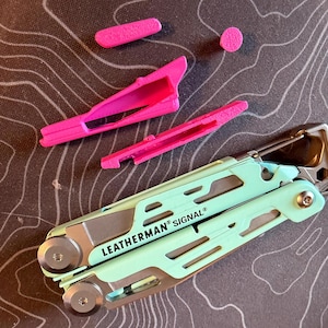 以下が含まれることがあります： ミントグリーンのハンドルとシルバーのアクセントが付いたマルチツールで、「LEATHERMAN SIGNAL」の文字が入っています。ピンク色のプラスチックアクセサリーが含まれています。ツールは、地形パターンを持つ暗い表面にあります。