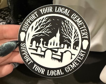 May include: A round black and white sticker with the text "SUPPORT YOUR LOCAL CEMETERY" encircling a cemetery scene. The scene includes a church, tombstones, and trees. The sticker is held by a hand with dark nail polish.