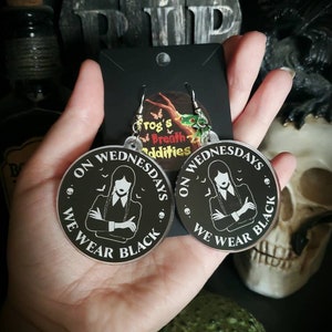 May include: A pair of black acrylic earrings with a white silhouette of Wednesday Addams from the Addams Family. The earrings feature the text "On Wednesdays We Wear Black" and a small bat graphic.