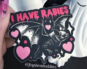 May include: A black sticker with a bat illustration, pink hearts, and the text "I HAVE RABIES" in dripping pink letters. The sticker also includes the text "@frogsbreathoddities".