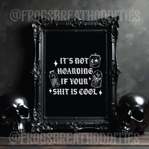 May include: A black framed print with the text "IT'S NOT HOARDING IF YOUR SHIT IS COOL" in white. The frame is flanked by two black skull sculptures. The print features illustrations of a jar, a scorpion in a bottle, and a terrarium.