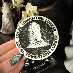 May include: A round sticker with the text "LOCAL TOMBSTONE LURKER" encircling a central illustration. The illustration features a tombstone with a draped figure and floral accents, all in black and white. The sticker is held in a hand with glittery blue nail polish.