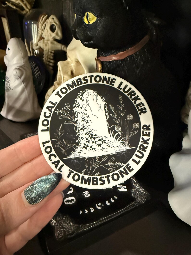 May include: A round sticker with the text "LOCAL TOMBSTONE LURKER" encircling a central illustration. The illustration features a tombstone with a draped figure and floral accents, all in black and white. The sticker is held in a hand with glittery blue nail polish.