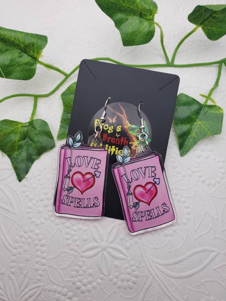 May include: A pair of pink acrylic earrings shaped like open books with the words "Love Spells" and a pink heart with an arrow through it.