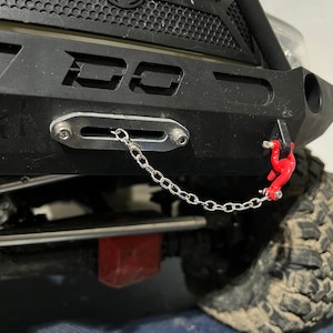 Puede incluir: Primer plano de un parachoques negro de vehículo todoterreno con una cadena plateada y un gancho rojo. El parachoques tiene una rejilla metálica y las letras "DD" recortadas. El neumático es visible en el lado derecho.