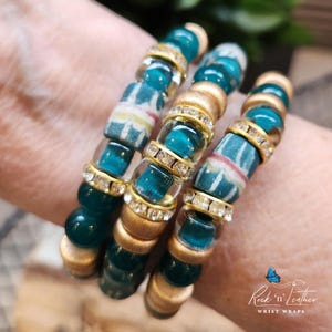 May include: Four stacked bracelets with teal and gold beads, some with clear rhinestones. The bracelets are on a person's wrist. The text "Rock 'n' Leather WRIST WRAPS" is visible at the bottom of the image.