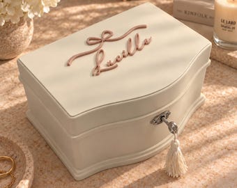 May include: A cream-colored rectangular jewelry box with a curved lid featuring the name Lucille in raised dusty pink script and a decorative bow. The box includes a silver-toned metal keyhole with a hanging white tassel key attached to the front.