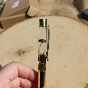 May include: A clear pen with a gold-colored metal barrel and clip. The pen's interior features a mushroom and moss encased in a clear resin. The pen is held over a wooden surface.