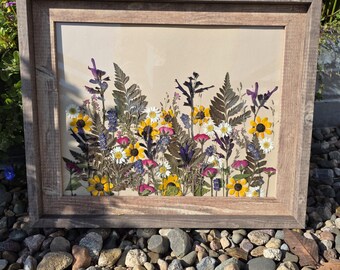 May include: A framed artwork featuring a pressed flower arrangement. The botanical display includes yellow, white, and purple wildflowers, along with green ferns, set against a beige background. The frame is made of weathered wood.