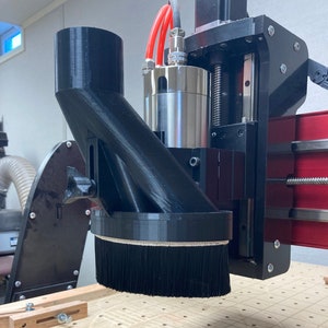 May include: A black dust collection system with a brush attachment, mounted on a CNC machine, over a wooden surface with a pattern carved into it.