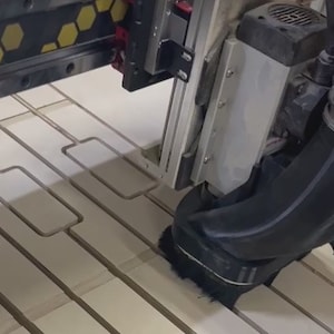 May include: A close-up of a CNC machine cutting out rectangular shapes from a piece of wood. The machine has a black dust collection system attached to it.