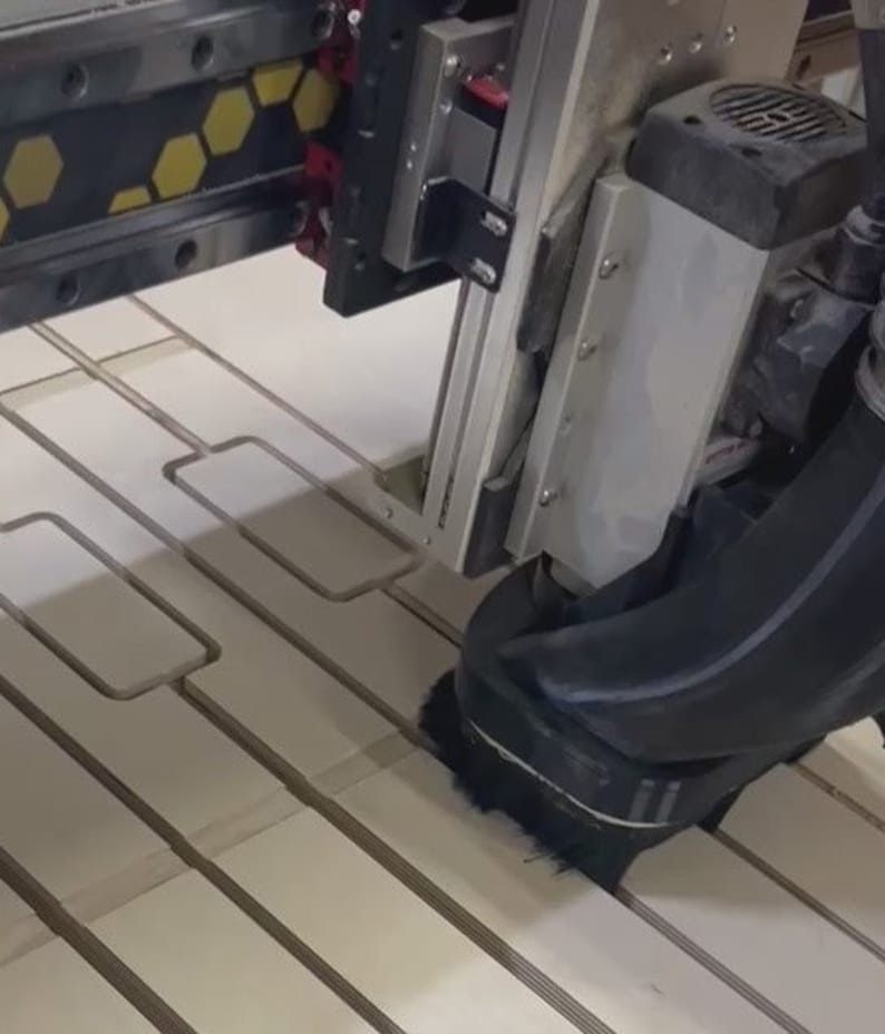 May include: A close-up of a CNC machine cutting out rectangular shapes from a piece of wood. The machine has a black dust collection system attached to it.