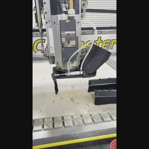 May include: A close-up of a CNC machine in operation. The machine has a silver and black color scheme, with a cutting tool in action. The machine is cutting into a light-colored surface, with a black brush nearby.