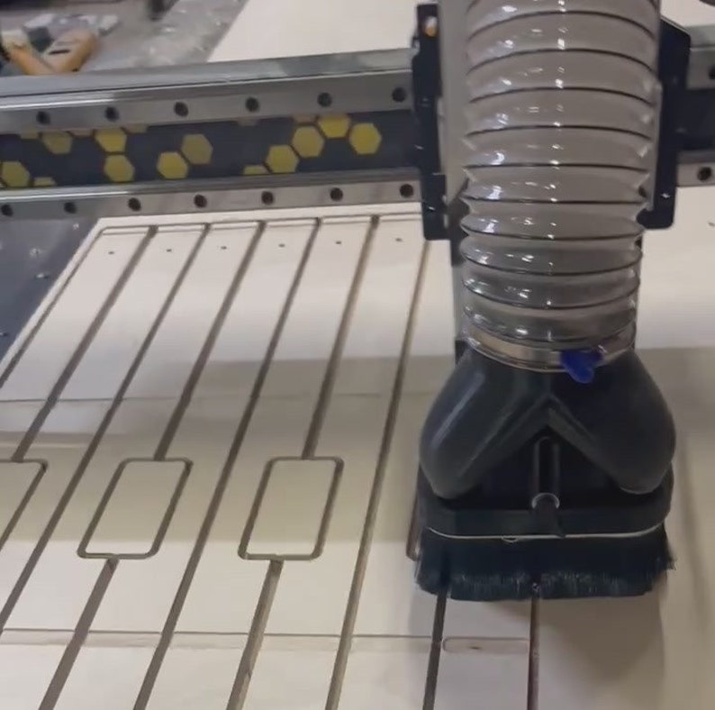 May include: A close-up of a CNC machine cutting out rectangular shapes from a piece of wood. A dust collection system is attached to the machine to remove sawdust.