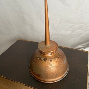 Puede incluir: Una aceitera de cobre vintage con un pico largo y c&oacute;nico. La lata tiene una base redondeada y una parte superior peque&ntilde;a y circular. Las palabras "MADE IN USA" est&aacute;n estampadas en el lateral. La aceitera est&aacute; sobre un libro marr&oacute;n oscuro.