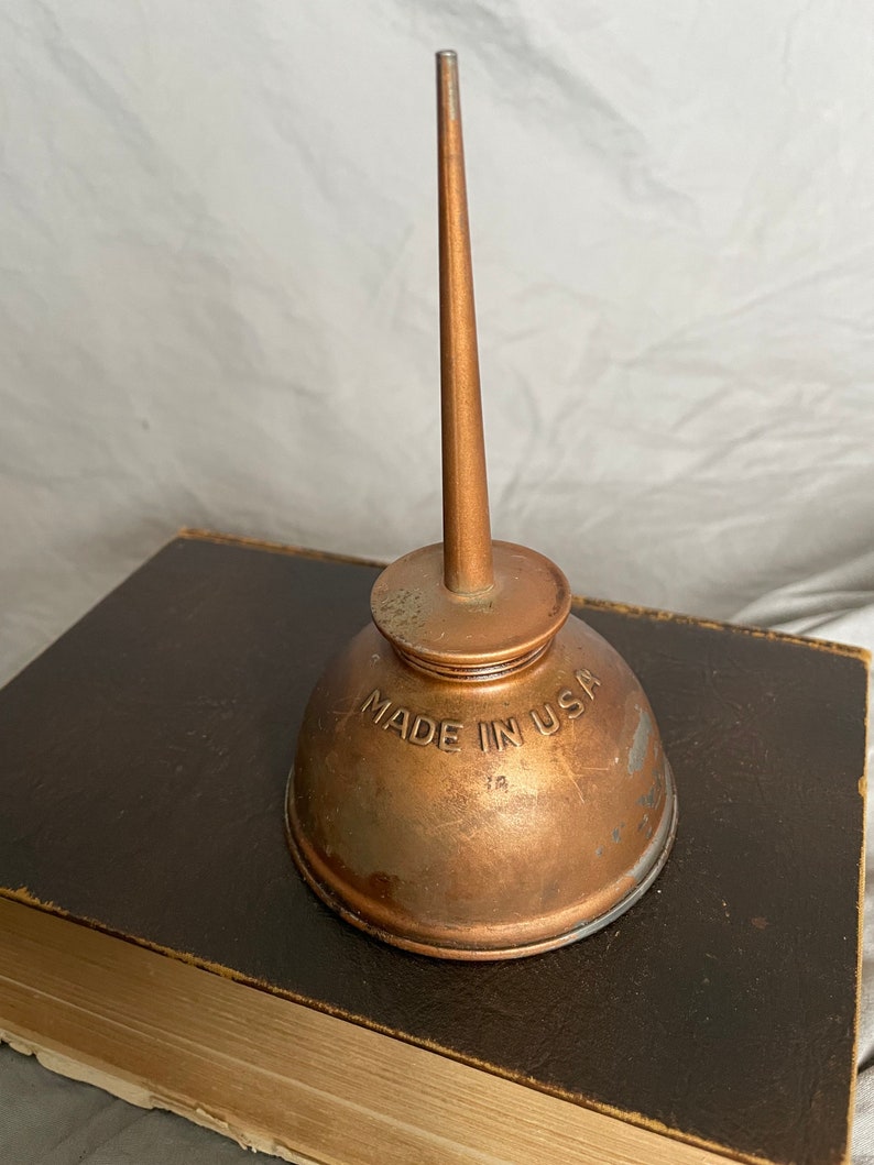 Puede incluir: Una aceitera de cobre vintage con un pico largo y c&oacute;nico. La lata tiene una base redondeada y una parte superior peque&ntilde;a y circular. Las palabras "MADE IN USA" est&aacute;n estampadas en el lateral. La aceitera est&aacute; sobre un libro marr&oacute;n oscuro.