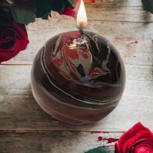 May include: A black and red marbled candle with a white wick is lit and burning. The candle is surrounded by red roses with green leaves.