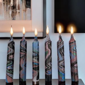 May include: Seven black, white, and blue marbled candles are lit and arranged on a dark brown surface. The candles are all the same size and shape, and they are all burning evenly.