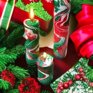 May include: Three red and green swirl candles with a white snowflake design on a red and green gift box. The candles are surrounded by green pine branches and red ribbon.