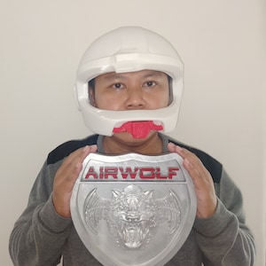 Puede incluir: Una persona con un casco blanco y una protección bucal roja sostiene un escudo plateado con la palabra "AIRWOLF" en letras rojas y un lobo con alas en el centro.