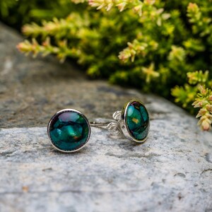 May include: A pair of silver stud earrings with iridescent green and blue abalone shell inlays. The earrings are on a grey stone surface.