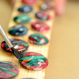 May include: A close-up of a hand using a paintbrush to apply a black sealant to a colourful round button. The button is on a wooden surface with other colourful buttons in a row.