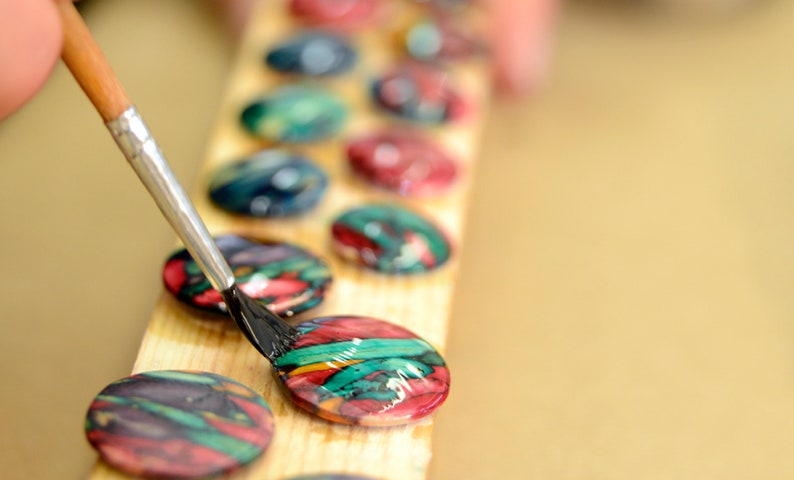 May include: A close-up of a hand using a paintbrush to apply a black sealant to a colourful round button. The button is on a wooden surface with other colourful buttons in a row.