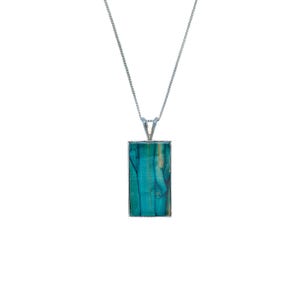 Puede incluir: Un collar plateado con un colgante rectangular. El colgante presenta un vibrante patrón turquesa y azul, enmarcado en plata. La cadena es delicada y cuelga de una fianza plateada.
