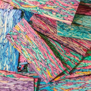 May include: A stack of colourful wooden blanks with a swirling pattern of pink, blue, green, and yellow. The wood is cut into rectangular shapes and is ready for crafting.