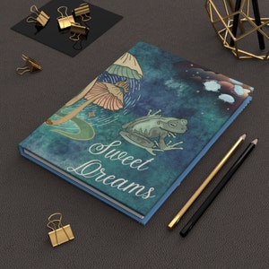 May include: A blue notebook with a watercolor background featuring a frog, mushrooms, and stars. The text "Sweet Dreams" is written in white cursive on the front cover.
