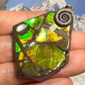 May include: A polished ammolite pendant featuring vibrant green and orange iridescent segments embedded in a dark matrix. The pendant is topped with a silver-toned metal bail shaped like a spiral ammonite fossil, showcasing a natural stone texture.