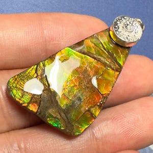 May include: A triangular pendant featuring a polished piece of iridescent ammolite gemstone with vibrant green, orange, and red color play, set in a silver-toned metal bail with a detailed spiral pattern engraved on the top surface.