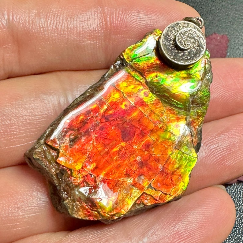 May include: A pendant featuring a polished, iridescent ammolite gemstone with vibrant red, orange, and green color shifts. The stone has a natural, irregular shape and is topped with a circular, textured silver-toned metal bail with a spiral design.