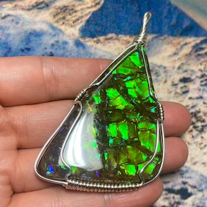 May include: A teardrop-shaped pendant featuring a polished ammolite gemstone with vibrant green and iridescent flashes, encased in a silver-colored wire-wrapped frame with a top loop for hanging, showcasing a mosaic-like surface texture.