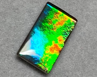 May include: A rectangular gemstone with a vibrant display of colors. The stone features a mix of green, orange, and blue hues, with a polished surface. The gemstone is set against a gray background.