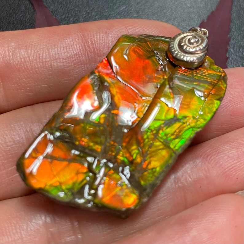 May include: A rectangular pendant featuring a polished piece of iridescent ammolite with vibrant orange, red, and green flashes across its surface. The gemstone is topped with a small, silver-toned metal bail shaped like a coiled ammonite fossil.