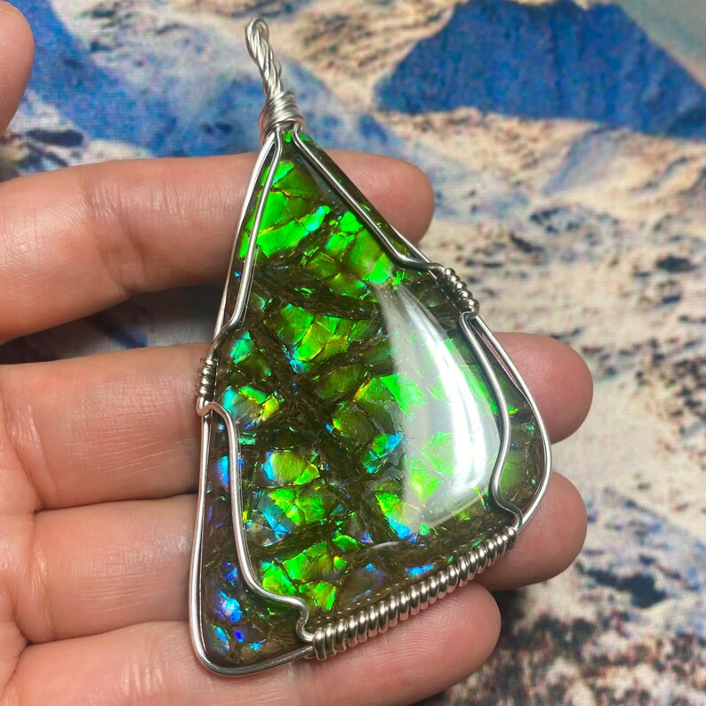 May include: A triangular pendant featuring a polished ammolite gemstone with vibrant green and blue iridescent patterns, encased in a silver-colored wire-wrapped frame with a twisted wire bail at the top for hanging on a necklace chain.